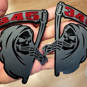 Könnte beinhalten: Zwei grau-schwarze Aufkleber zum Thema Sensenmann. Jeder zeigt einen Schädel mit einer Sense und die rote Zahl "345". Die Aufkleber werden in einer Hand gehalten, um ihr Design und ihre Details zu zeigen.