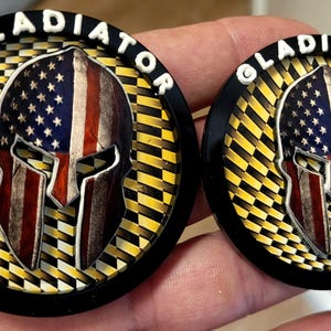 May include: Two round black Gladiator emblems. Each emblem features a Spartan helmet design with the American flag incorporated into the helmet's design. The word "GLADIATOR" is in white, raised lettering around the edge of each emblem.