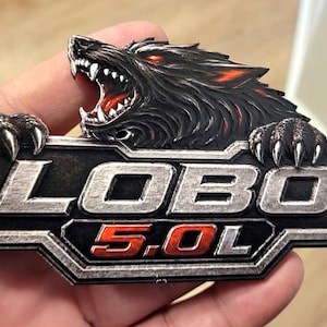 May include: A black and silver Lobo 5.0L emblem featuring a snarling wolf with red eyes and accents. The emblem has a rectangular shape with the word "LOBO" and "5.0L" in a bold font.