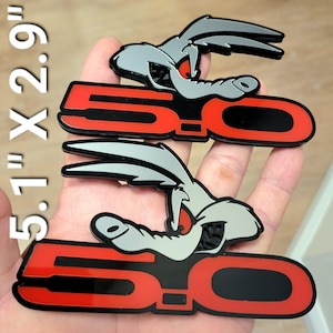 Puede incluir: Dos emblemas de coche negros, rojos y grises con un conejo de dibujos animados y el número "5.0". Cada emblema mide 13 cm x 7,4 cm. El conejo tiene orejas y cara grises. El "5.0" es rojo con contorno negro.