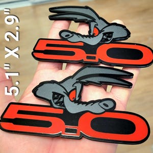 May include: Two black and red 5.0 emblems with a cartoon coyote design. Each emblem measures 5.1" x 2.9". The coyote has gray fur, red eyes, and a menacing expression. The "5.0" text is in red with a black center.