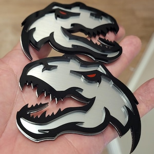 Può includere: Due emblemi a forma di testa di dinosauro. Gli emblemi hanno una faccia color argento con contorni neri e denti affilati. L'occhio del dinosauro è rosso. Gli emblemi sono tenuti nella mano di una persona.