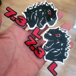 Puede incluir: Dos pegatinas de Godzilla en blanco y negro con ojos rojos y el texto "7.3" en rojo. Las pegatinas presentan una ilustración detallada de la cabeza de Godzilla con dientes afilados y una aleta dorsal puntiaguda.