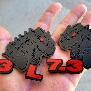 Puede incluir: Dos emblemas automotrices negros y rojos con una cabeza de dinosaurio con ojos rojos. El texto "7.3 L" es rojo. Los emblemas son probablemente para un vehículo.