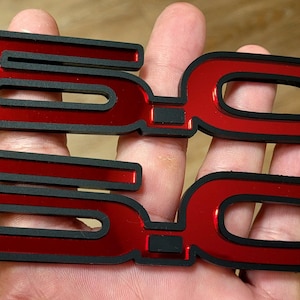 Puede incluir: Dos emblemas 5.0 rojos y negros. Los emblemas tienen un acabado rojo brillante con un contorno negro. Los números están estilizados y parecen estar hechos de un material reflectante. Los emblemas están en la mano de una persona.