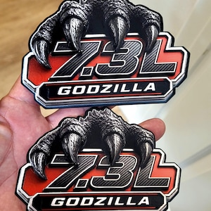 以下が含まれることがあります： 黒とシルバーの7.3Lゴジラエンブレムステッカー2枚。各ステッカーは、「7.3L」の文字と白い「GODZILLA」の文字の上にモンスターの爪のデザインが施されています。背景は赤と黒です。