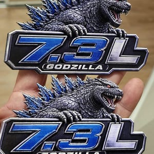 May include: Two Godzilla-themed emblems. Each emblem features a detailed, gray Godzilla figure with blue dorsal fins and red eyes, positioned above a blue and silver emblem that reads "7.3L GODZILLA".