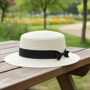 May include: A cream-coloured straw hat with a black ribbon and bow rests on a wooden picnic table. The hat features a flat top and brim, with a black ribbon tied in a bow. The background shows a park with trees and a path.