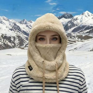May include: A beige fleece hood with a built-in face covering and drawstring. The hood is worn over a striped shirt. The background features snow-covered mountains.