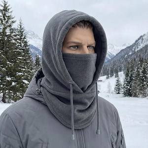 May include: A gray fleece balaclava with a hood and face covering. The balaclava has a drawstring and is worn over a gray jacket. The background shows a snowy mountain landscape.