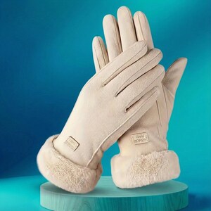 Può includere: Un paio di guanti beige con una morbida texture simile alla pelle scamosciata. I guanti hanno un polsino in ecopelliccia e una piccola etichetta rettangolare. I guanti sono esposti su un piedistallo turchese su sfondo blu.