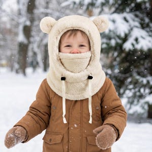 May include: A child wearing a cream-colored, fuzzy winter hat with bear ears and a matching neck warmer. The hat has a drawstring and black toggles. The child is also wearing a brown coat and mittens, with a snowy background.