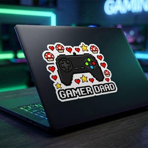 May include: A black laptop featuring a pixel art sticker on its lid. The sticker displays a black game controller surrounded by red hearts, yellow stars, and red-and-white mushrooms, with the text GAMER DAAD printed in white pixelated letters.