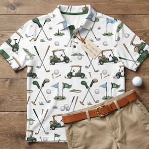 Può includere: Polo bianco con un motivo a tema golf con golf cart, mazze, palline e bandiere. Pantaloncini color cachi e una cintura marrone sono accanto alla camicia. È visibile anche una pallina da golf. L'etichetta dice "Happy Father's Day".
