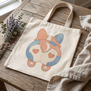 Puede incluir: Una bolsa de tela color crema con un gráfico central de la parte trasera de un corgi. La ilustración muestra una paleta de colores melocotón y azul con dos pequeños corazones rojos en los cuartos traseros y huellas visibles abajo.