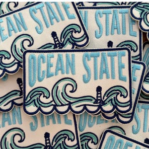 May include: A rectangular embroidered patch featuring the text OCEAN STATE in light blue thread. The design includes stylized teal and dark blue ocean waves and a small lighthouse icon, all bordered by a dark blue stitched edge on a white background.