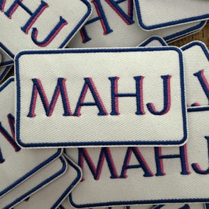 May include: A rectangular embroidered patch featuring the text MAHJ in dark blue letters with a pink shadow effect. The patch has a white background and a dark blue stitched border, presented as a collection of multiple identical patches.