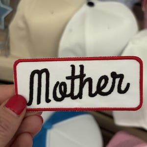 May include: A rectangular embroidered patch with a white background and a red border. The word Mother is stitched in black cursive script across the center of the patch. The patch has rounded corners and a clean, finished edge design.