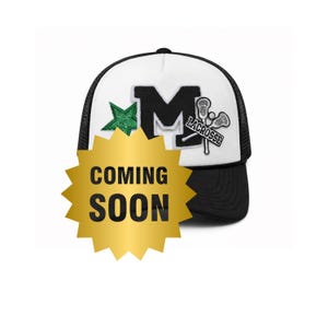 Puede incluir: Gorra de camionero en blanco y negro con un panel frontal blanco con una gran "M" negra y una estrella verde. La gorra también tiene un gráfico de lacrosse y las palabras "COMING SOON" en una estrella dorada.