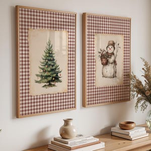 Pode incluir: Duas impressões de arte de Natal emolduradas. Uma apresenta uma árvore de Natal em aquarela, a outra um boneco de neve segurando uma cesta. Ambas estão sobre um fundo xadrez vermelho e branco e emolduradas em madeira.