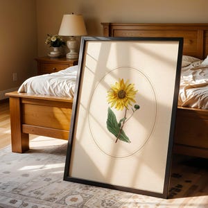 May include: A framed botanical print featuring a yellow sunflower with green leaves and a bud, set against a cream-colored background within an oval. The black frame leans against a bed in a sunlit bedroom.