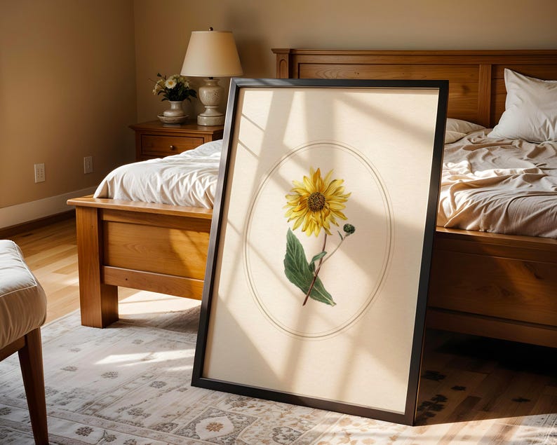 May include: A framed botanical print featuring a yellow sunflower with green leaves and a bud, set against a cream-colored background within an oval. The black frame leans against a bed in a sunlit bedroom.