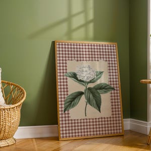 May include: A framed botanical print of a white hydrangea flower with green leaves. The artwork is set against a cream background and framed with a brown and white gingham pattern. The frame is a light wood colour.