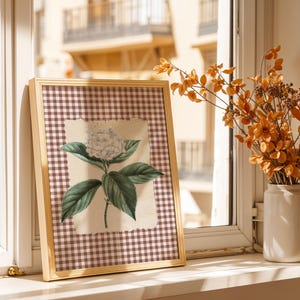 May include: A framed botanical print of a white hydrangea flower and green leaves. The print is set against a brown and white checked background and framed in light wood. A vase of orange flowers sits nearby.