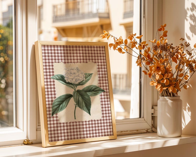 May include: A framed botanical print of a white hydrangea flower and green leaves. The print is set against a brown and white checked background and framed in light wood. A vase of orange flowers sits nearby.