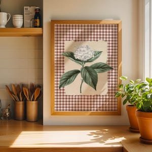 May include: A framed botanical print of a white hydrangea flower with green leaves. The artwork is set against a brown and white gingham background and framed in light wood. Two potted plants sit on a wooden countertop.