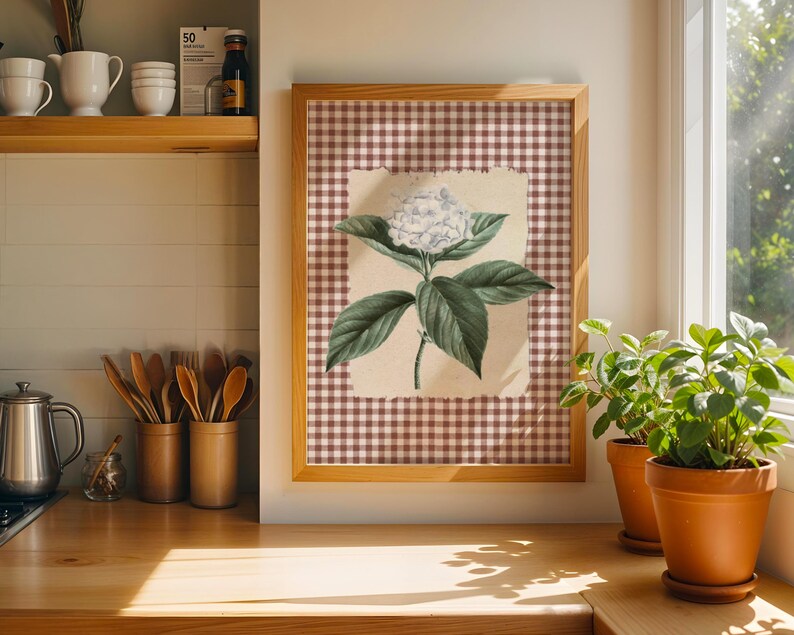 May include: A framed botanical print of a white hydrangea flower with green leaves. The artwork is set against a brown and white gingham background and framed in light wood. Two potted plants sit on a wooden countertop.