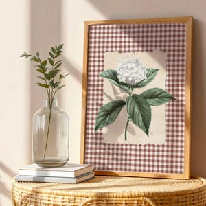 May include: A framed botanical print of a white hydrangea flower with green leaves, set against a brown and white checked background. A clear glass vase with greenery and two books are on a wicker table.