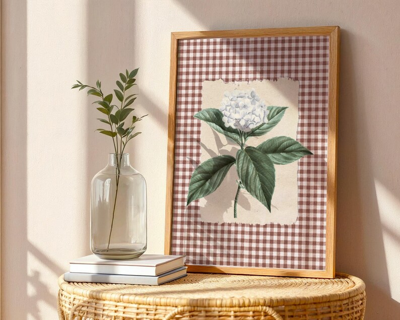 May include: A framed botanical print of a white hydrangea flower with green leaves, set against a brown and white checked background. A clear glass vase with greenery and two books are on a wicker table.