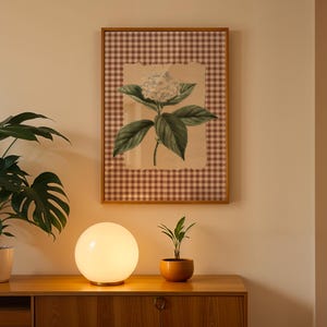 May include: A framed botanical print of a hydrangea flower with green leaves and a white bloom, set against a brown and white checkered background. A glowing orb lamp sits on a wooden cabinet, with a potted plant on the left and a small plant in an orange pot on the right.