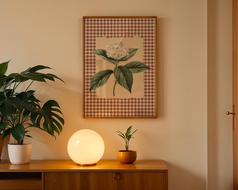 May include: A framed botanical print of a hydrangea flower with green leaves and a white bloom, set against a brown and white checkered background. A glowing orb lamp sits on a wooden cabinet, with a potted plant on the left and a small plant in an orange pot on the right.