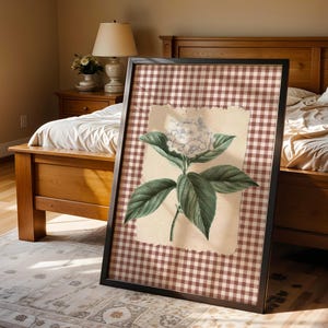 May include: Framed botanical print of a white hydrangea flower with green leaves. The artwork is set against a tan background and a brown and white gingham pattern. The black framed print is displayed in a bedroom setting.