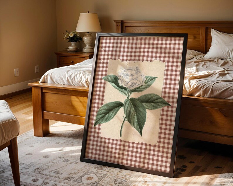May include: Framed botanical print of a white hydrangea flower with green leaves. The artwork is set against a tan background and a brown and white gingham pattern. The black framed print is displayed in a bedroom setting.