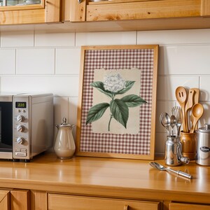 May include: A kitchen setting with a framed botanical print of a white hydrangea flower and green leaves. A stainless steel microwave, a creamer, and kitchen utensils are on a wooden countertop. White tiles and wooden cabinets are in the background.