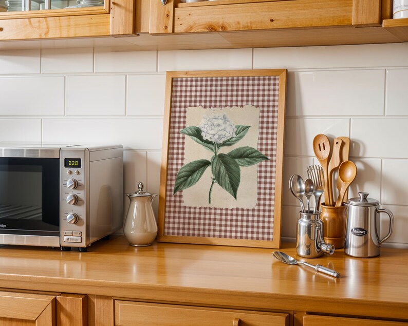 May include: A kitchen setting with a framed botanical print of a white hydrangea flower and green leaves. A stainless steel microwave, a creamer, and kitchen utensils are on a wooden countertop. White tiles and wooden cabinets are in the background.