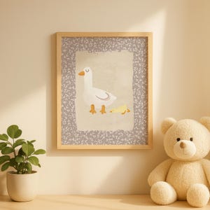May include: A framed print featuring a whimsical illustration of a white goose with orange beak and feet, and a yellow duckling. The artwork is set against a light beige background with a gray floral border, encased in a light wood frame.