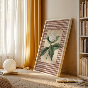 May include: A framed botanical print with a white flower and green leaves on a checkered background. The artwork is in a white frame and leans against a wall near a bookshelf and window with yellow curtains. A white orb sits on a book.