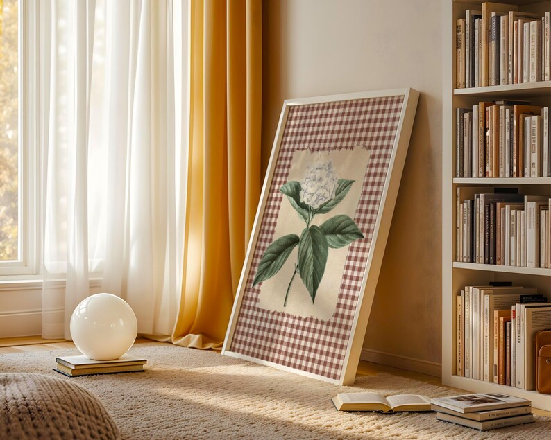 May include: A framed botanical print with a white flower and green leaves on a checkered background. The artwork is in a white frame and leans against a wall near a bookshelf and window with yellow curtains. A white orb sits on a book.