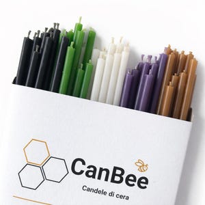 Può includere: Una scatola di candele colorate in varie tonalità, tra cui nero, verde, bianco, viola e marrone. Le candele sono disposte in una scatola bianca con il logo "CanBee" e la scritta "Candele di cera".