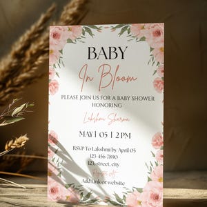 May include: A baby shower invitation with a floral design. The text reads "BABY In Bloom" with event details. The invitation is framed with pink flowers and green leaves. The event is for Lakshmi Sharma on May 1st at 12 PM.