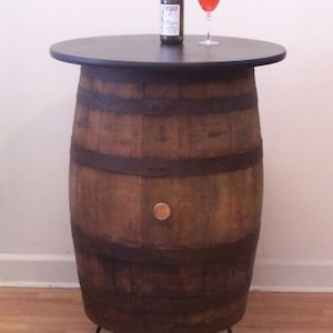 May include: A wooden barrel table with a black top and metal legs. The table is about 30 inches tall and has a diameter of about 20 inches. A bottle of wine and a glass of wine are on the table.