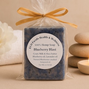 May include: A blue soap bar, labelled "Blueberry Blast", is wrapped in cellophane and tied with a raffia bow. The soap is next to a white flower, a stack of stones, and a lit candle in a small holder. The label lists ingredients, with a net weight of 107g.
