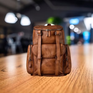 May include: A brown leather backpack with multiple compartments and straps. The backpack has a top flap with a zipper closure and two side pockets with zippers. The bag is sitting on a wooden surface.