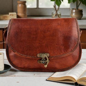May include: A brown leather satchel bag with a curved flap and a metal clasp. The bag has a shoulder strap and is sitting on a white surface. The bag is a medium size and appears to be new.
