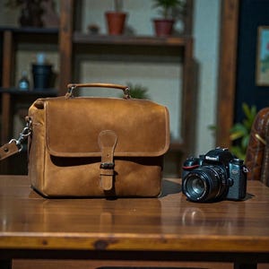 May include: A brown leather camera bag with a top handle, buckle closure, and shoulder strap sits on a wooden table. A black Nikon camera with a lens is next to the bag. The bag and camera are in a studio setting.