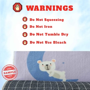 Puede incluir: Una alfombra de color morado claro con un gr&aacute;fico de un oso blanco sobre una luna creciente azul. El texto incluye WARNINGS, Do Not Squeezing, Do Not Iron, Do Not Tumble Dry y Do Not Use Bleach, con iconos de stop rojos y manos.